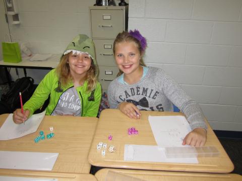 Two smiling students, one dressed in a dragon costume, work on an assignment at their desks